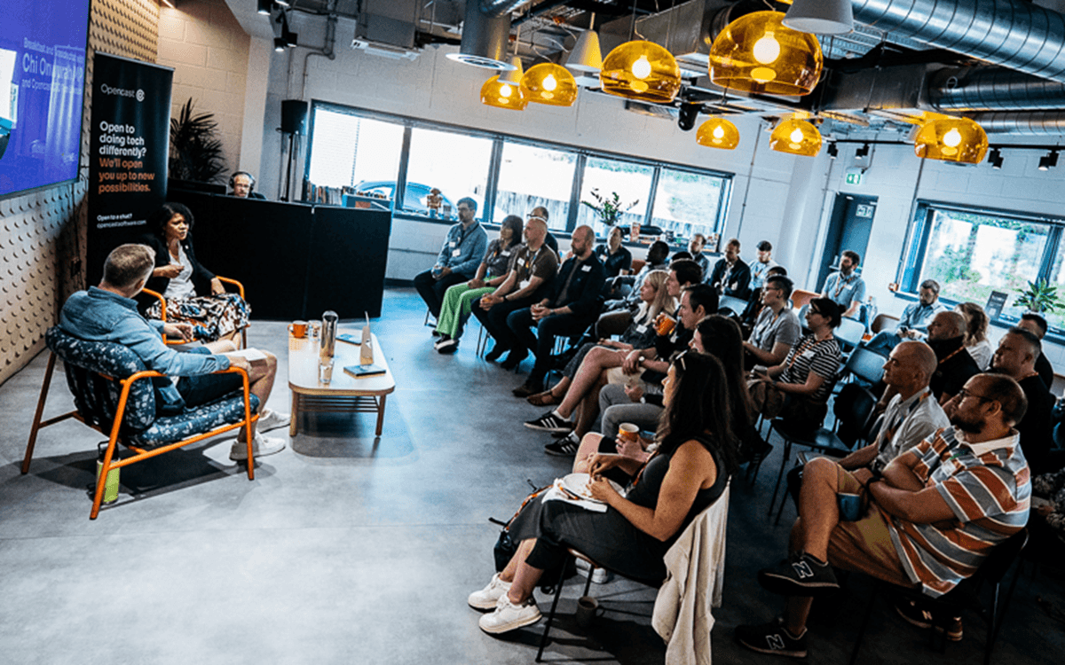 A wide view of an audience attending a talk or panel discussion in a modern, industrial‑style indoor venue. Rows of seated attendees face a small stage area where two people are seated in conversation. Warm pendant lights hang from the ceiling, and large windows along one side of the room flood the space with natural light. Event signage and a screen are visible near the front, creating the atmosphere of a professional talk, meetup, or community discussion.