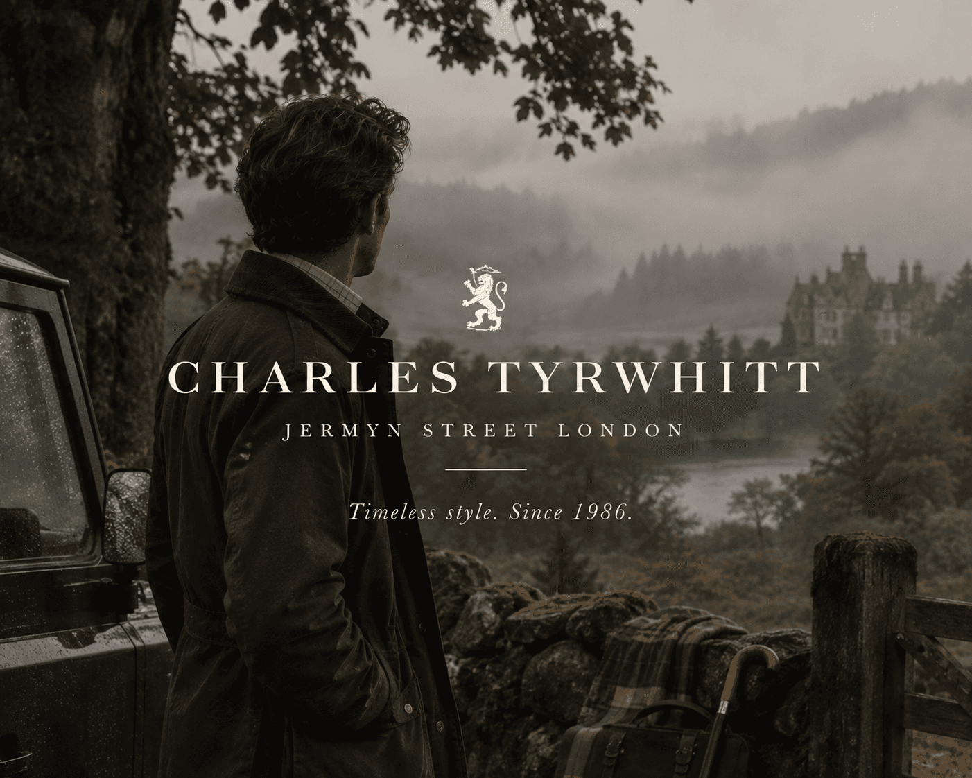 A back profile view of a man wearing a dark olive field jacket standing beside a vintage off-road vehicle near a stone wall. He looks out over a misty, atmospheric Scottish Highlands landscape featuring a lake, pine trees, and a distant castle estate. In the foreground rests a leather holdall bag, a tartan plaid blanket, and a classic wooden umbrella. The center overlay features white brand text with a lion crest reading "CHARLES TYRWHITT JERMYN STREET LONDON" and the tagline "Timeless style. Since 1986."