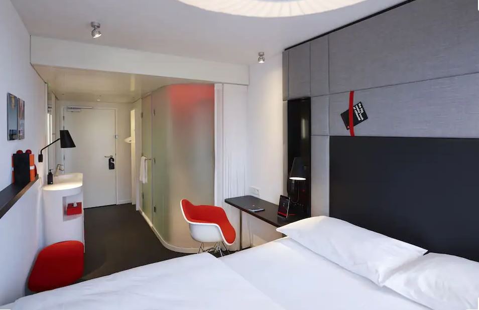 Modern hotel room interior featuring an oversized bed, a small desk and chair, and stylish red accents woven throughout.