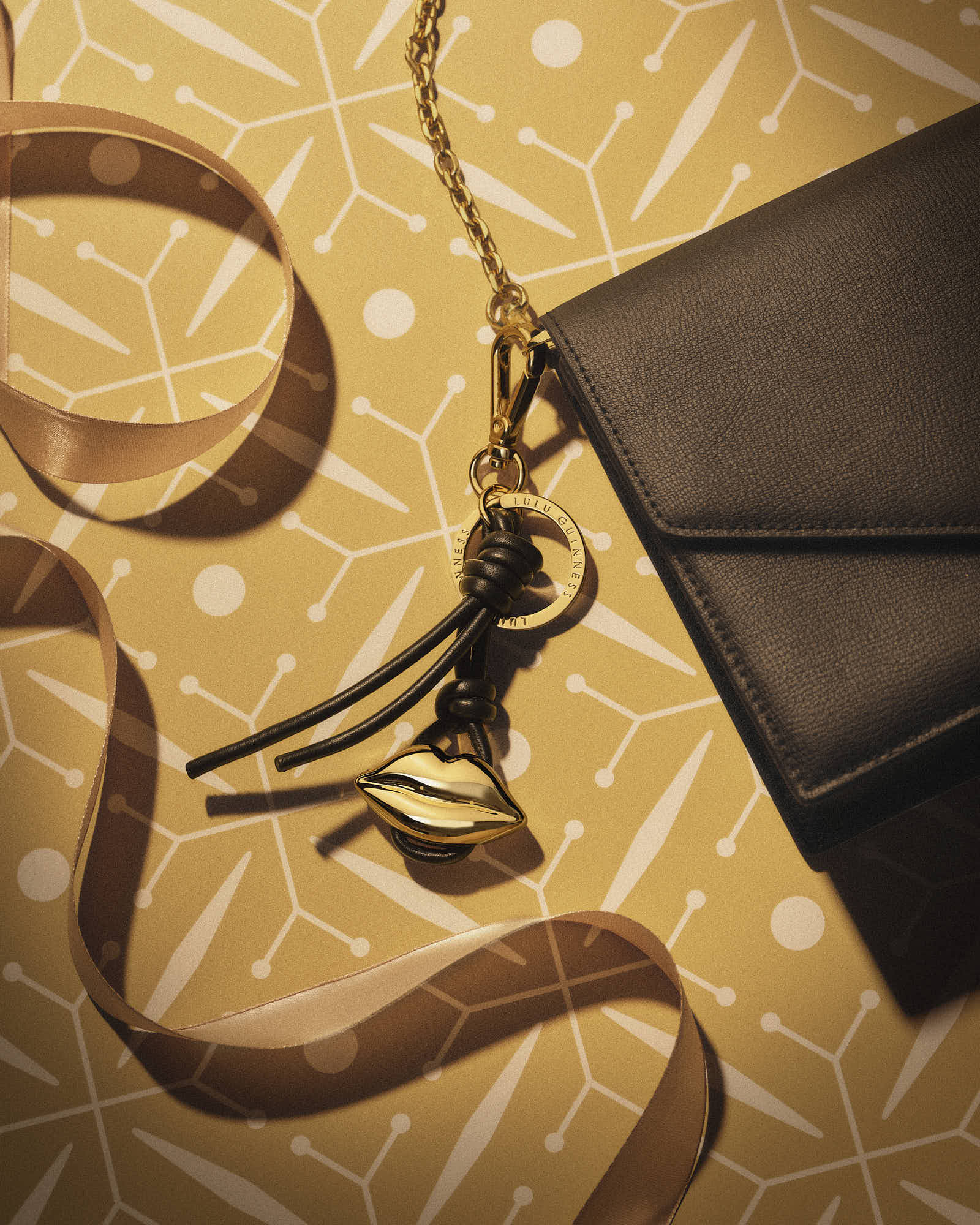 Black handbag with gold lips charm and ribbon on patterned background.