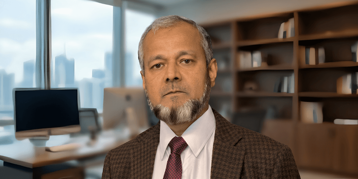 An older man with a salt-and-pepper beard wears a suit and tie, standing in a modern office with large windows, bookshelves, and computer monitors.