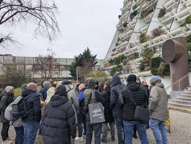 A group of people in winter clothing stand outside next to a uniquely designed building with plants on its balconies.