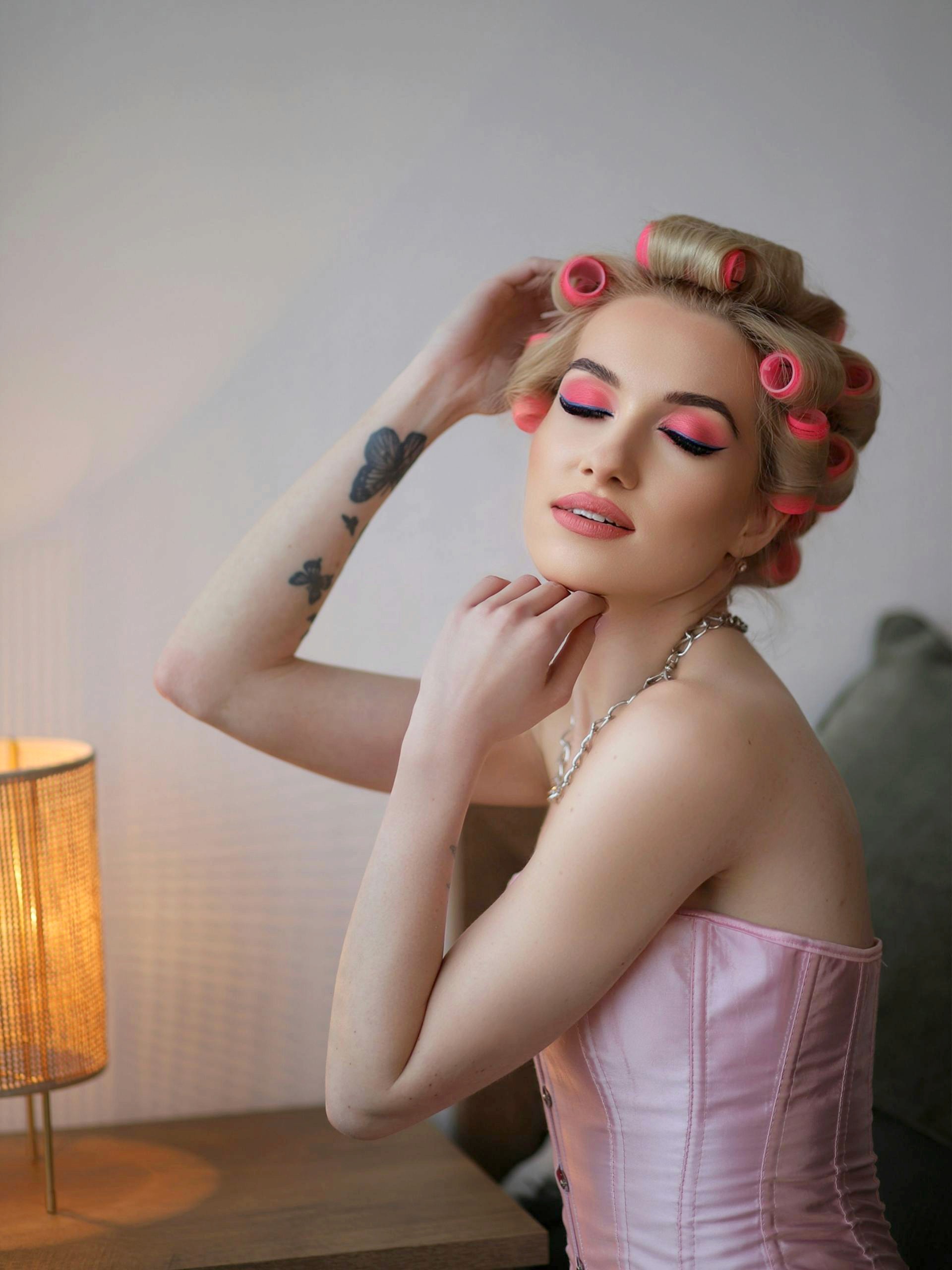 A woman with blonde hair in pink rollers and vibrant pink eye makeup posing with her eyes closed, wearing a pink corset and silver chain necklace.