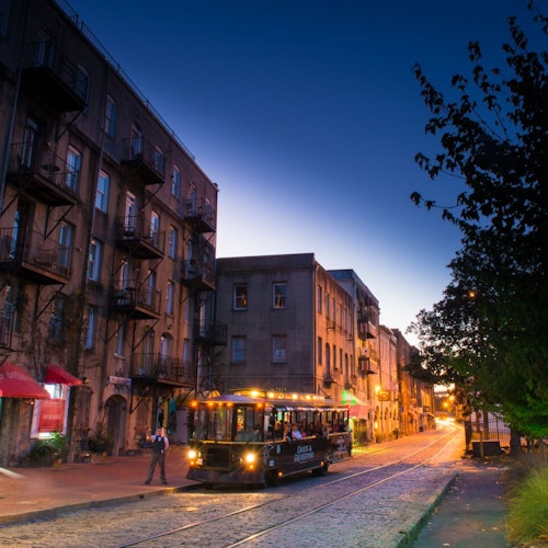 傍晚时分,一辆有轨电车沿着鹅卵石街道行驶,街道两旁是古老的建筑和灯火通明的商店。人行道旁种满了树木。