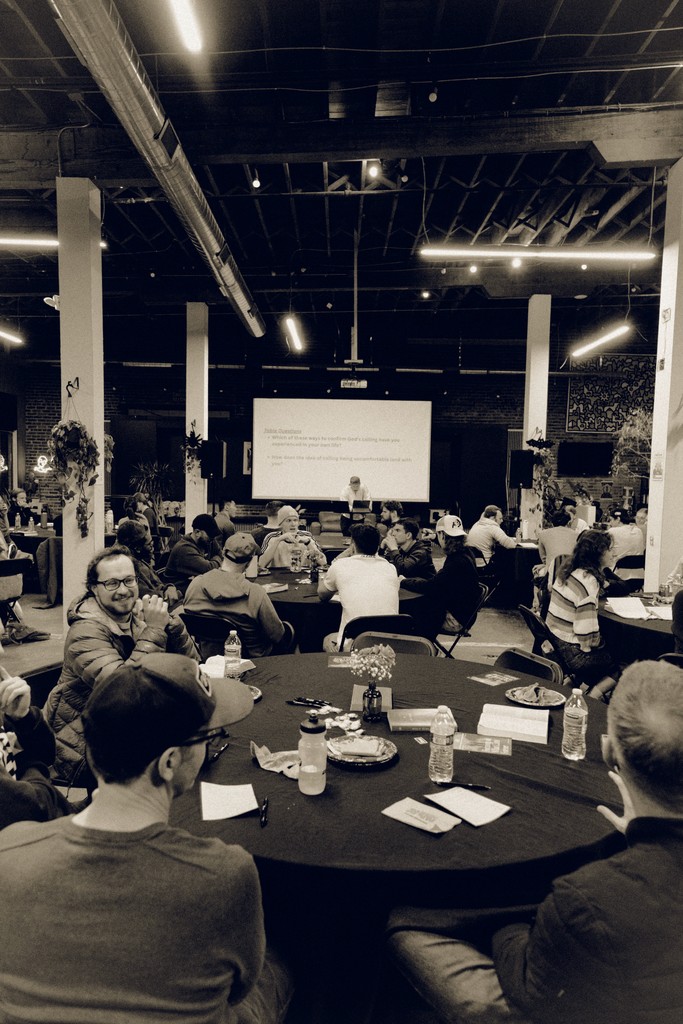 Room full of people at roundtables conversating, connecting, and sharing stories. Speaker standing in front of room preaching.