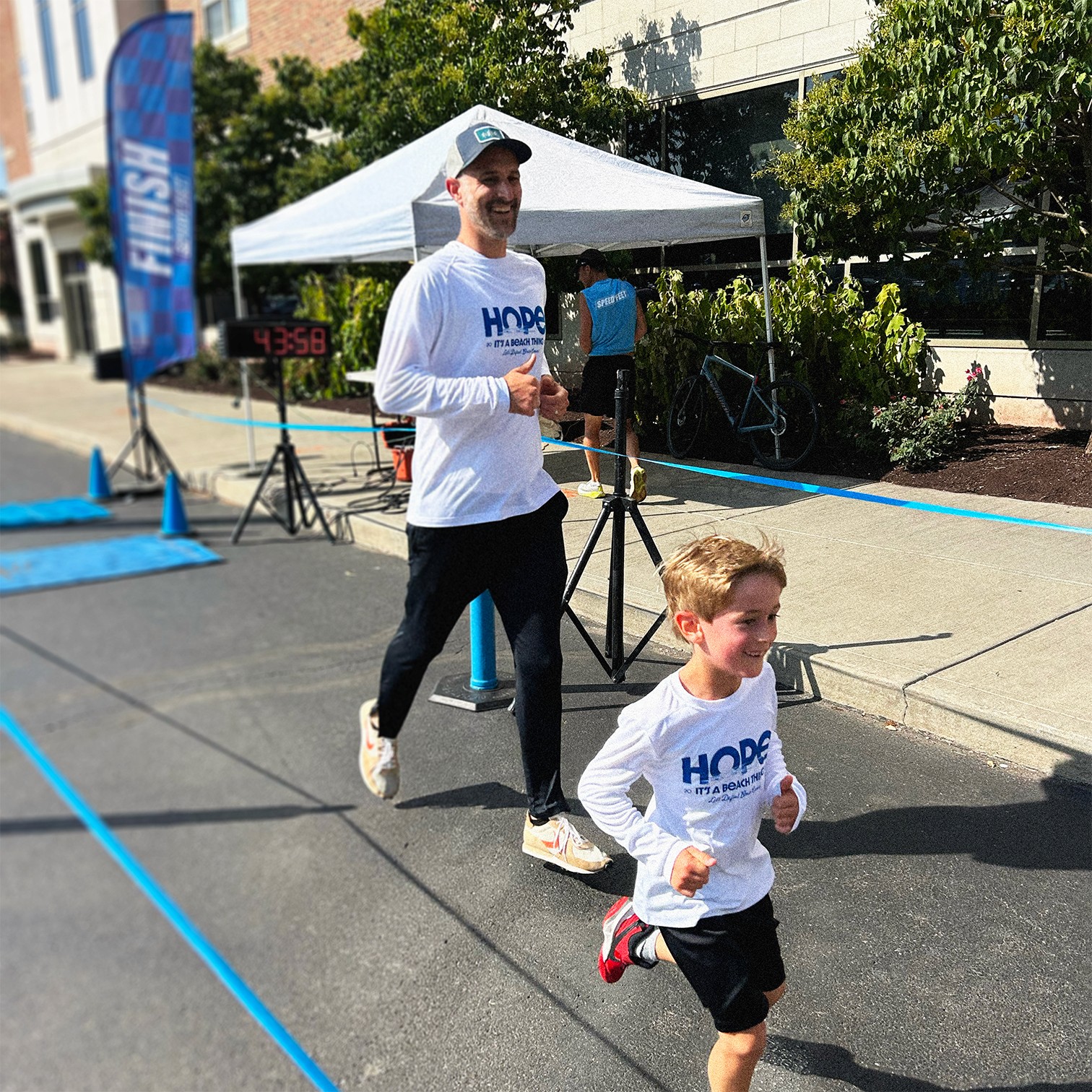 Man and boy finishing the 'HOPE IT'S A BEACH THING' 5K marathon, wearing long-sleeved white 'HOPE IT'S A BEACH THING' branded shirts.