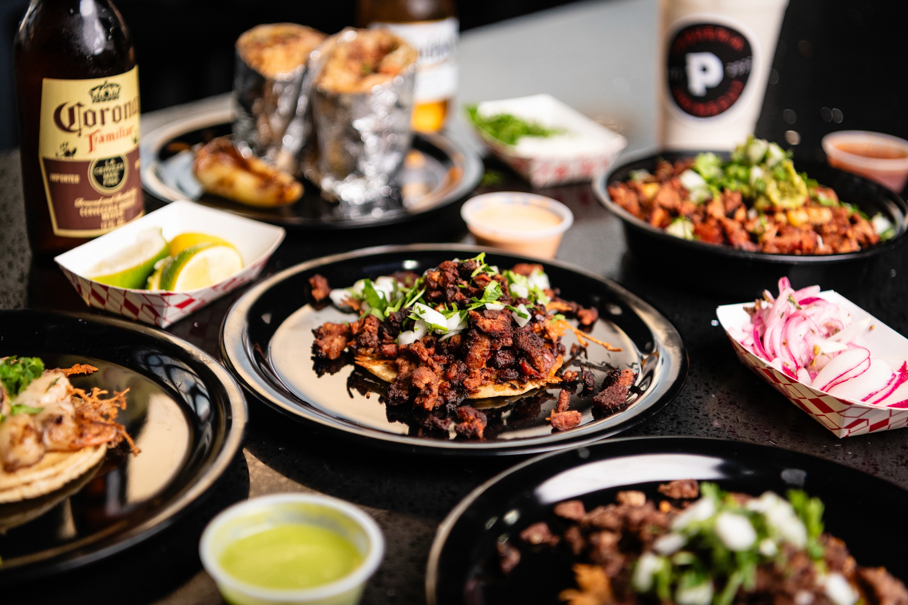 Grilled tacos topped with onion and cilantro, foil-wrapped burritos, salsas, pickled onions, and drinks on a Primecut Taqueria table.
