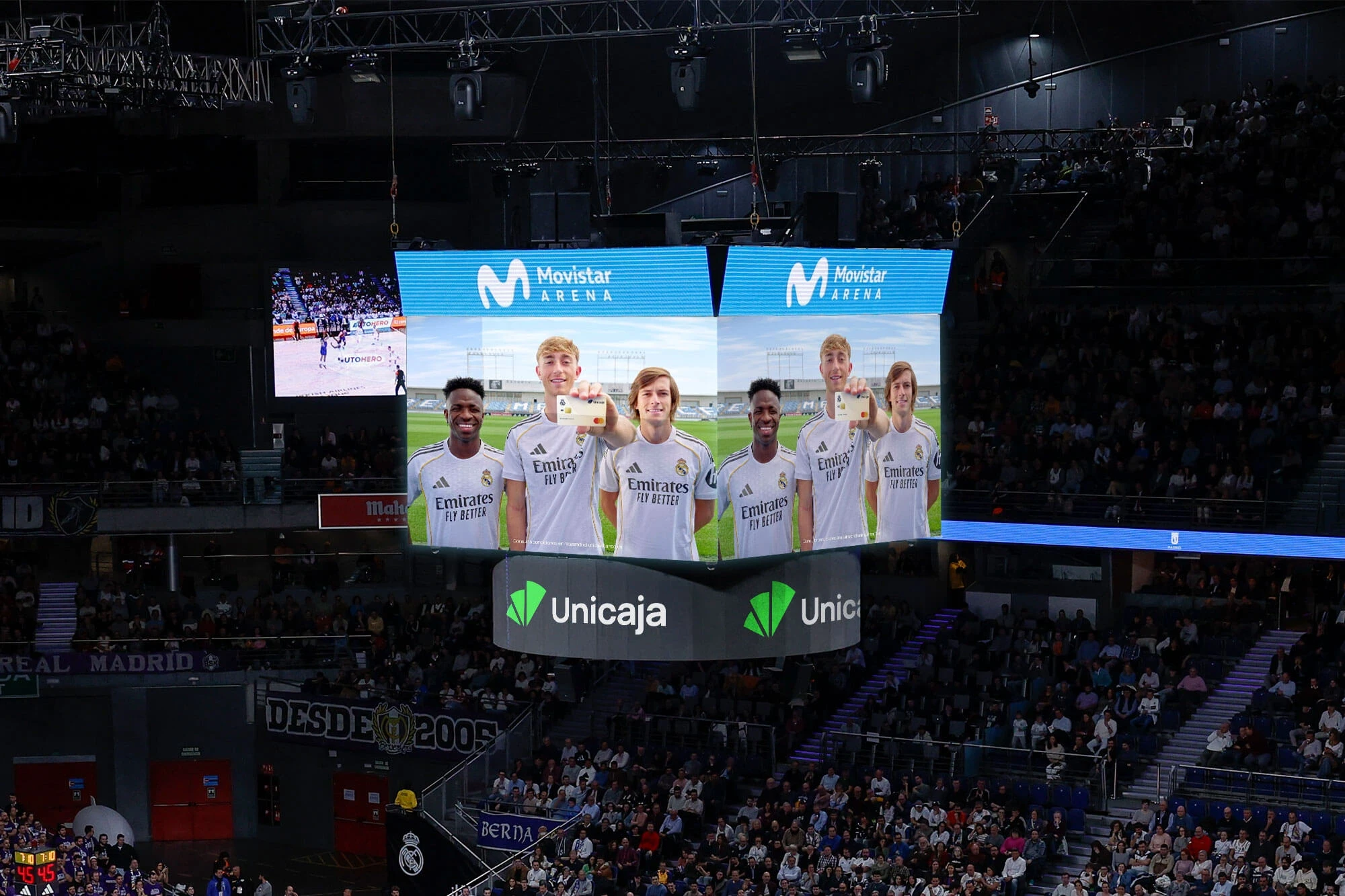 Pantalla gigante en el interior de un pabellón deportivo proyectando la campaña publicitaria de la tarjeta con jugadores del primer equipo
