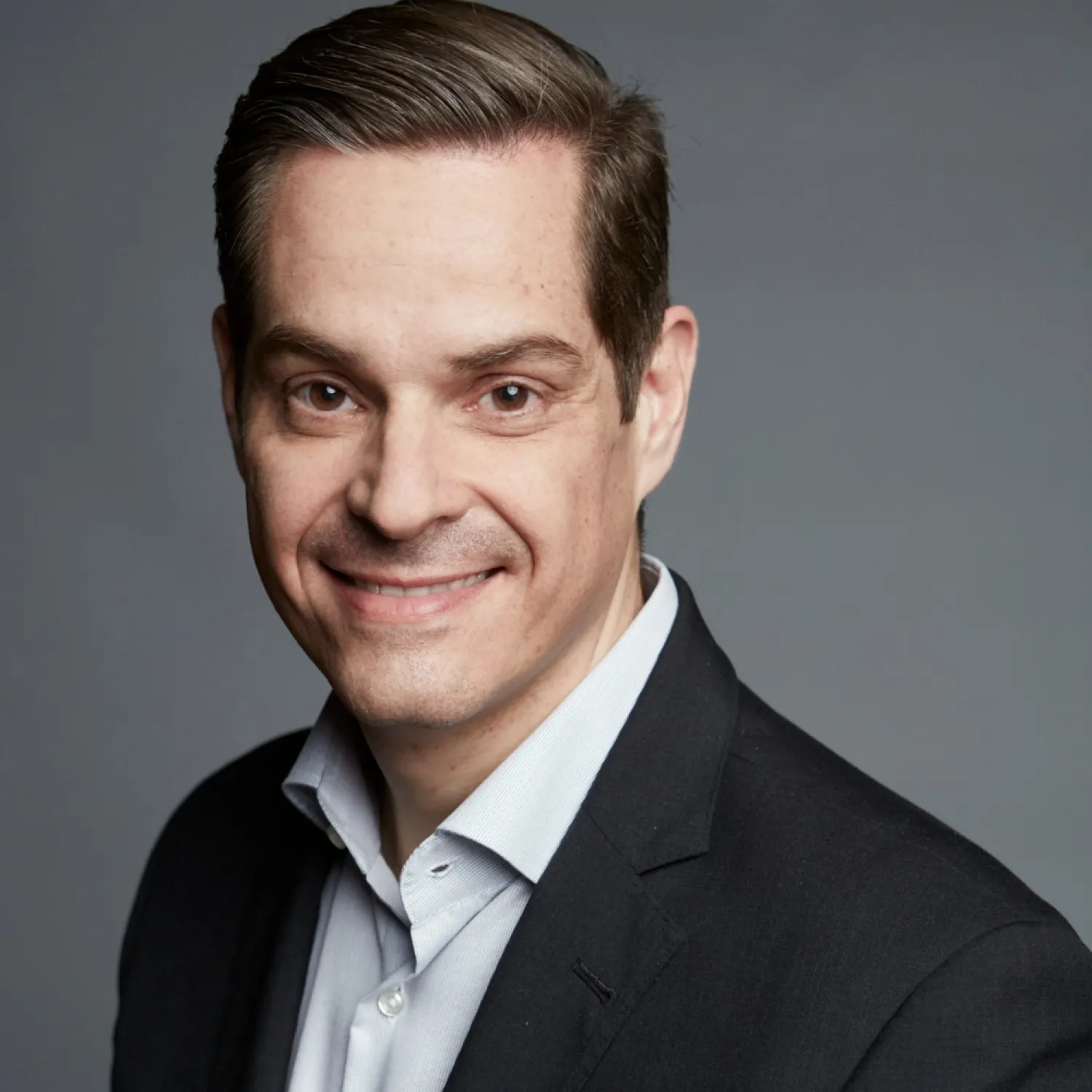 A young person with curly hair smiles at the camera while sitting in front of a laptop against a dark background.