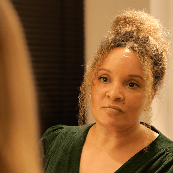 A woman with tousled brown hair wears round orange sunglasses and a floral-patterned blouse. Her expression is neutral, conveying a vintage vibe.