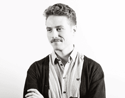 A black and white portrait of a man with short, slightly curly hair and a mustache, looking off to his left. He is wearing a light-colored collared shirt under a dark cardigan or sweater. His arms are crossed over his chest. The background is plain white.