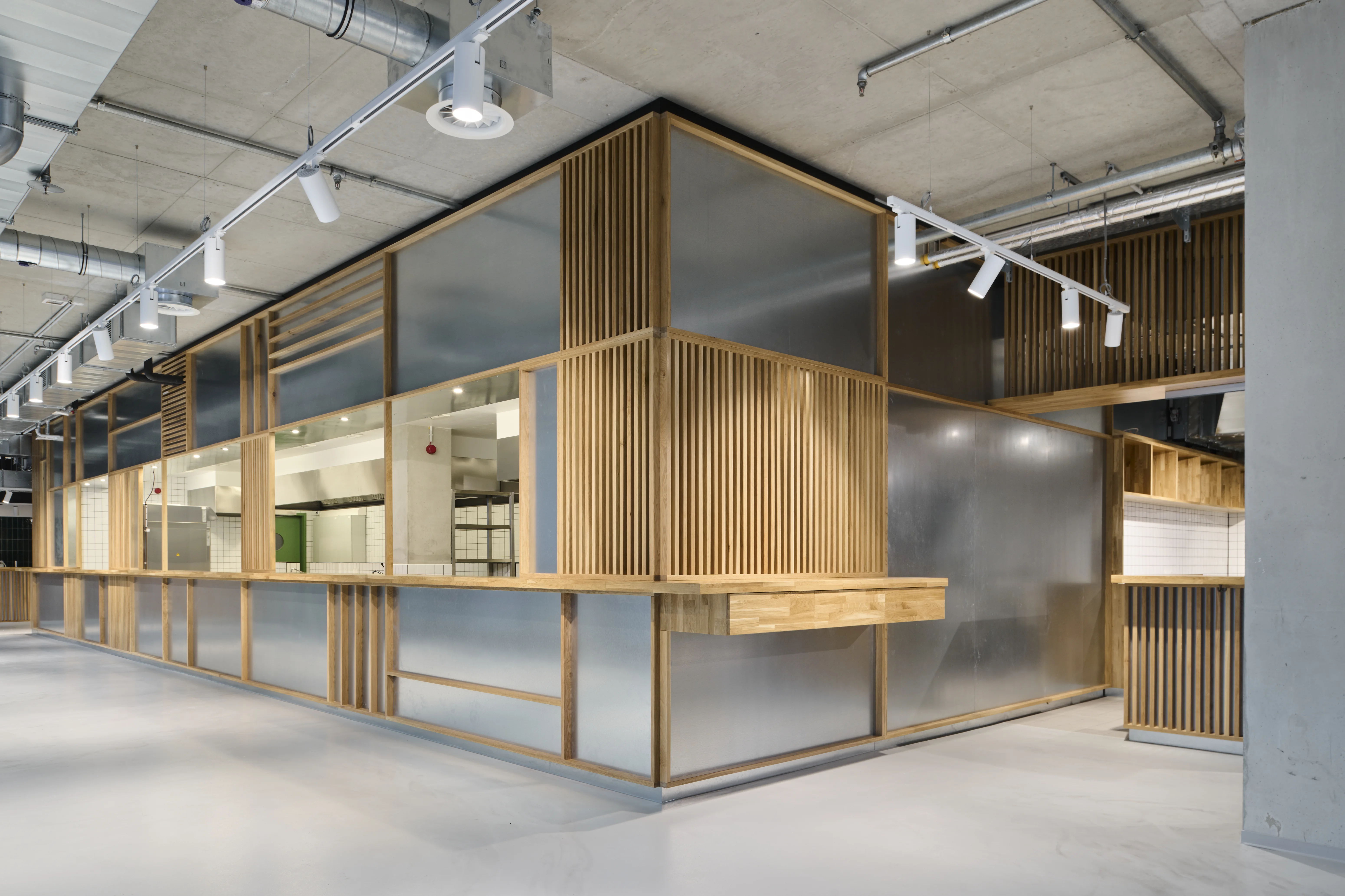Open kitchen cube at PARKER BOWLES & FRIENDS framed in wooden slats and frosted glass panels under an exposed concrete ceiling with industrial lighting.