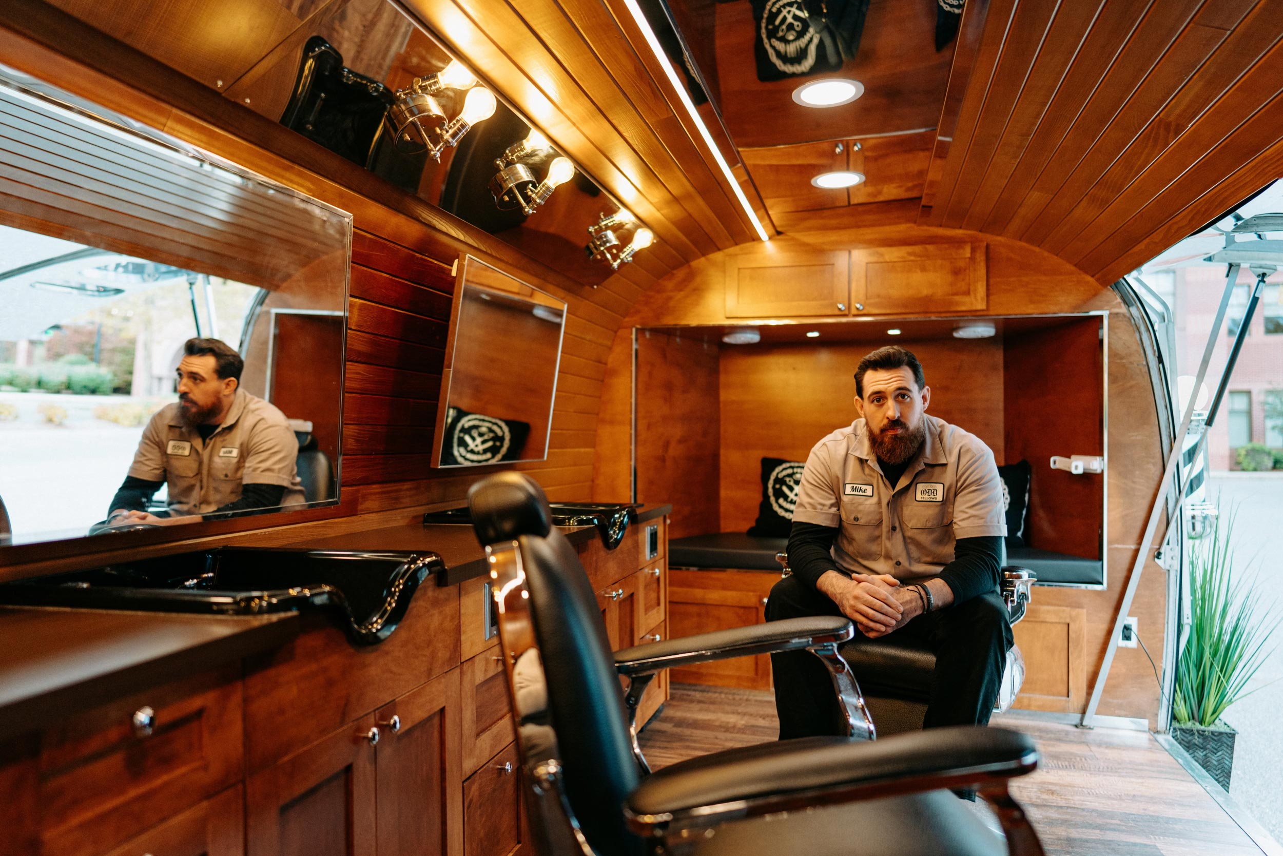 A man with a full beard and a tan work shirt is sitting on a cushioned bench inside a customized mobile barbershop. The interior features rich, dark wood paneling, recessed lighting, and a black leather barber chair in the foreground with wash basins and vanity mirrors. The man is looking directly at the camera.