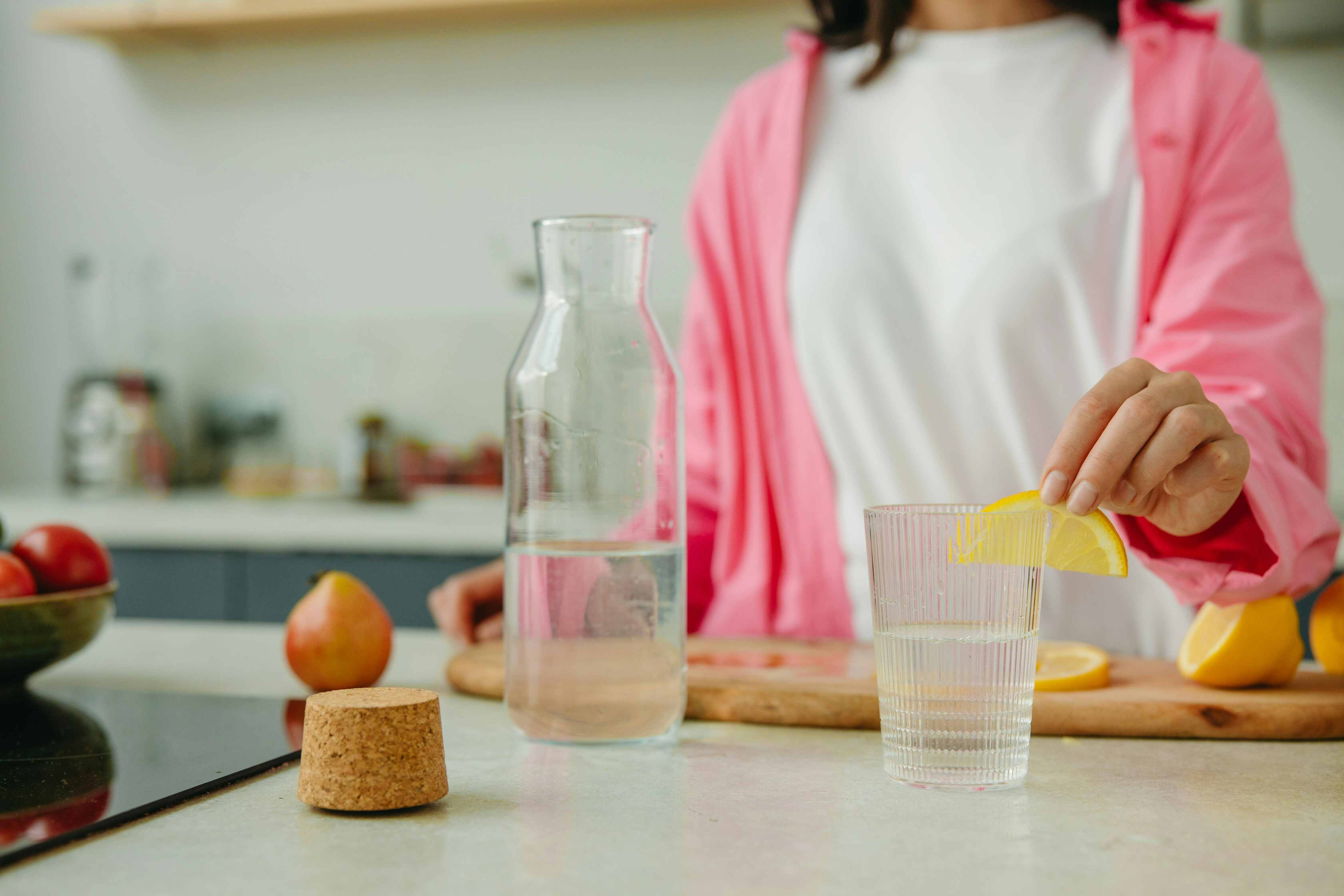A woman in a pink jacket adds a lemon slice to water, promoting hydration, digestion, and metabolic health in a bright kitchen.