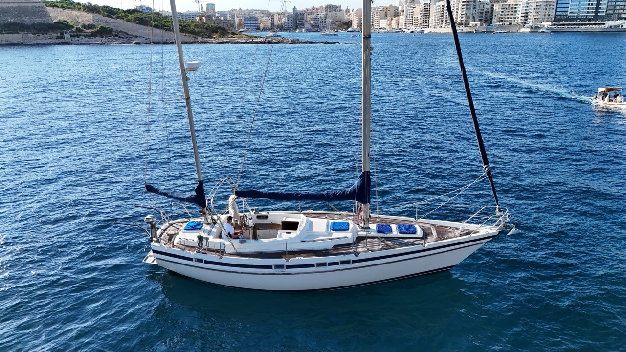 Contest 38 Ketch motoring through Marsamxett Harbour Malta under power with sails furled — classic white offshore ketch with teak deck and twin masts, Sliema waterfront and Valletta bastions visible in the background