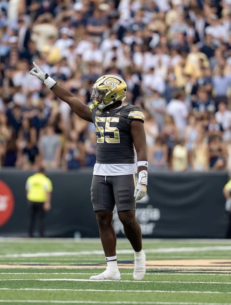 Christian Garrett in a gray and gold uniform with the number 55 stands on a field, pointing with his right arm raised. A crowd watches in the background.