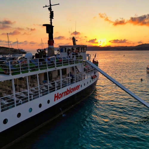 Човен під назвою "Hornblower" на воді під час заходу сонця з людьми на палубі, віддаленими пагорбами та помаранчевим небом.
