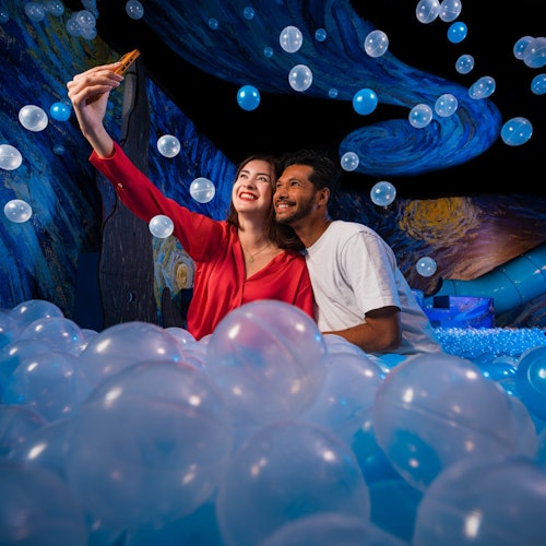 Una pareja se toma una selfie mientras está sentada en una piscina llena de pelotas de plástico azules y blancas, rodeada de remolinos artísticos azules.