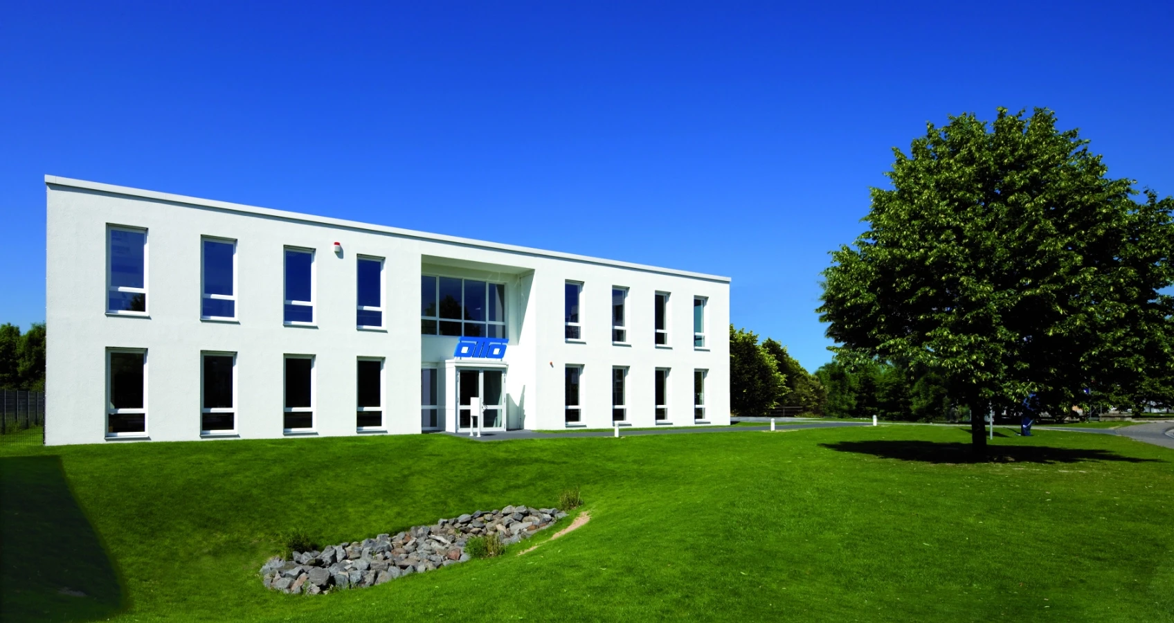 Modernes weißes Bürogebäude mit vielen Fenstern, umrahmt von einem gepflegten Rasen und einem großen Baum unter blauem Himmel.
