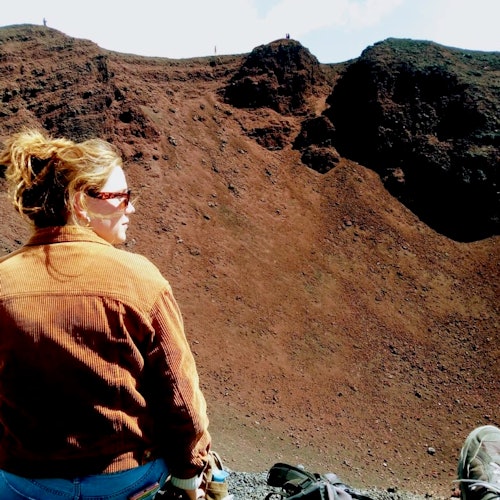 Pessoa com óculos de sol e casaco castanho está em frente a uma encosta rochosa, de cor vermelho-acastanhada, sob um céu parcialmente nublado.