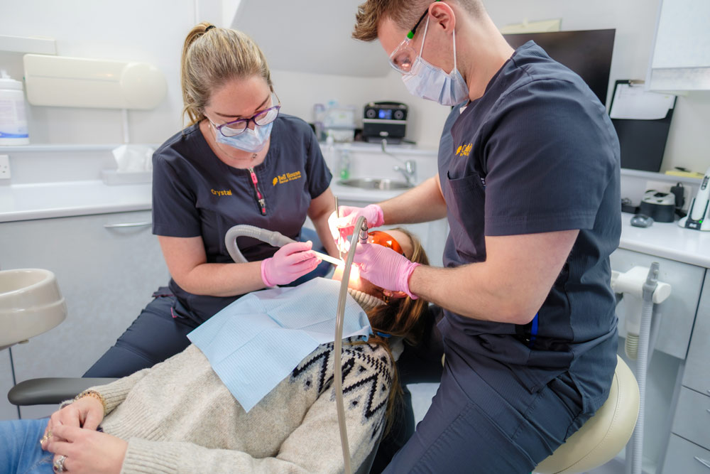Two dental professionals in gray scrubs, wearing masks and gloves, are performing a dental procedure on a patient in a modern clinic, with dental equipment and a monitor visible in the background.