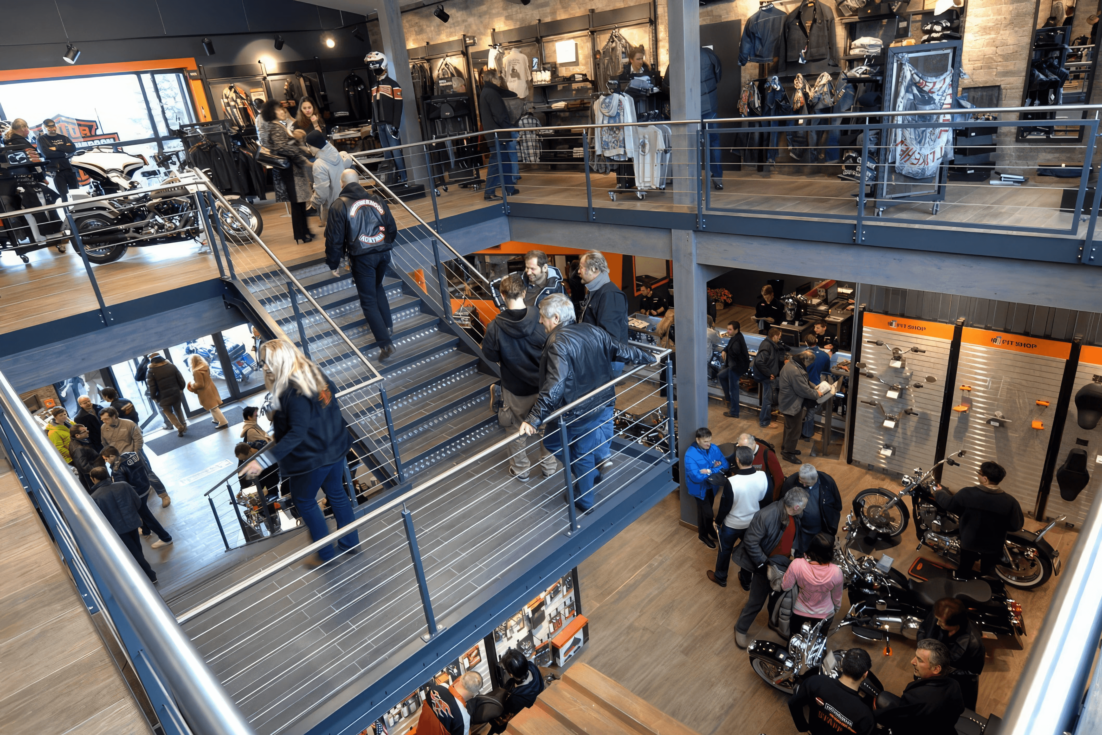 MW Interior  - Belebter zweigeschossiger Harley-Davidson Motorrad-Showroom mit Stahltreppe, Motorrädern auf beiden Ebenen, Zubehörwänden und zahlreichen Besuchern bei Eröffnung" Beschreibung: Atmosphärischer Blick in den Motodrom Harley-Davidson Showroom bei der Eröffnung – zwei Ebenen verbunden durch eine Stahltreppe, Motorräder in Szene gesetzt auf Holzparkett, Zubehör- und Bekleidungswände auf beiden Etagen und ein belebtes Publikum, das die neue Fläche in Besitz nimmt.