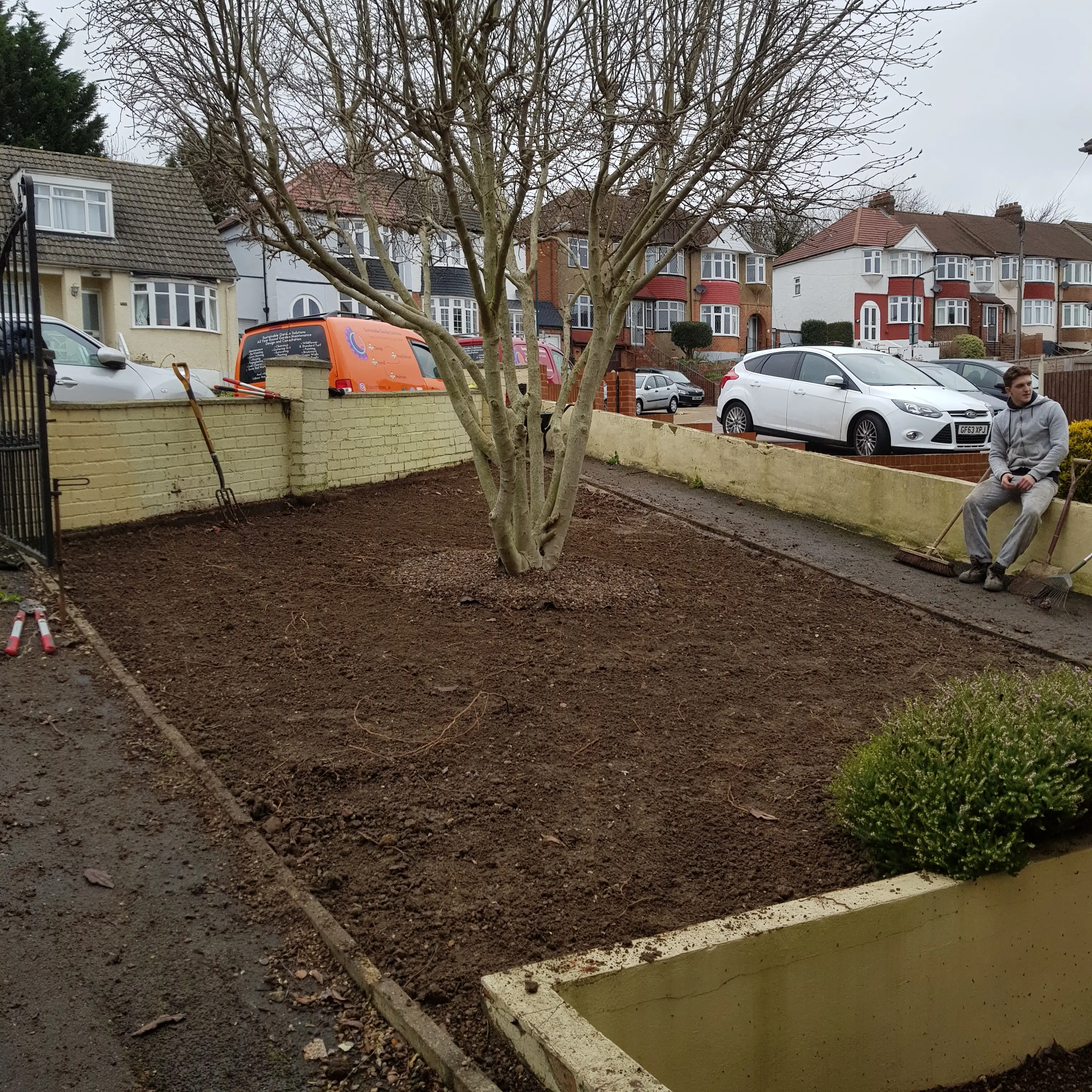 A garden plot with a tree, surrounded by mulch, and structures in the background. People are visible nearby.