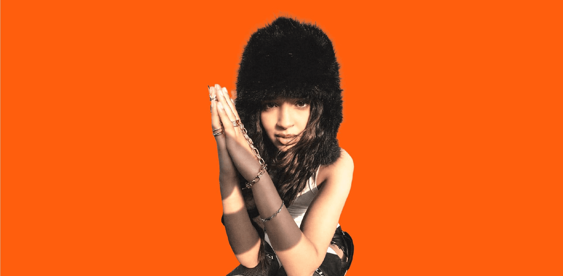 Bannière au format panoramique avec un fond uni orange vif (couleur d'identité de TopLine). À droite, un portrait tronqué et stylisé d'une artiste musicale (rap/pop) ou d'une influenceuse. Elle porte un bonnet en fausse fourrure noire et de multiples bagues et chaînes dorées. Ses mains sont jointes devant son visage, créant une pose accrocheuse. L'espace négatif à gauche est dédié au texte. Ce visuel de direction artistique forte est utilisé pour l'identité visuelle et le branding du média digital musical TopLine, mettant en évidence les services de graphisme et de création de contenu de Nux-Studio.