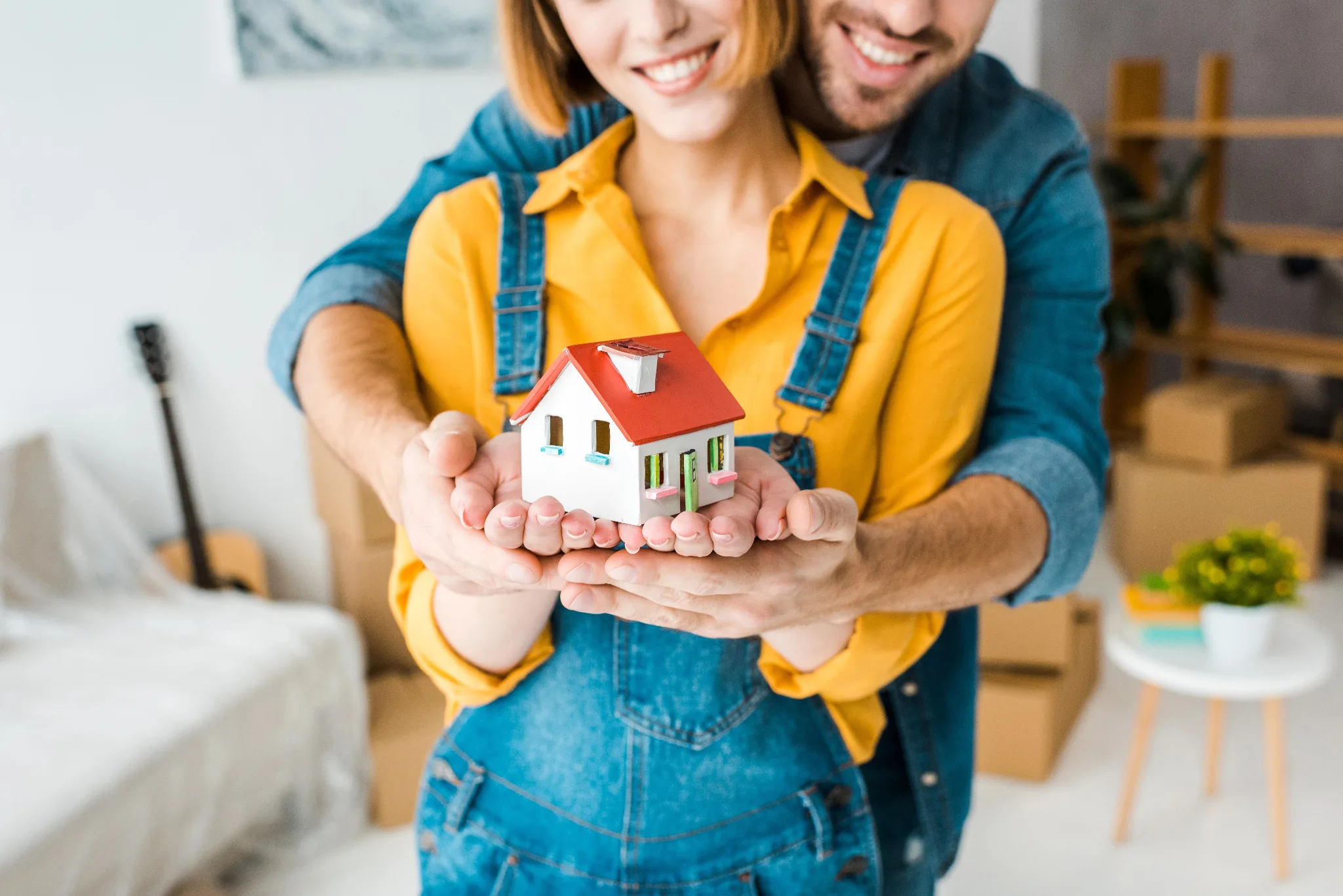 A man standing behind his wife, who is wearing a yellow shirt and overalls, both holding a miniature house in the palms.