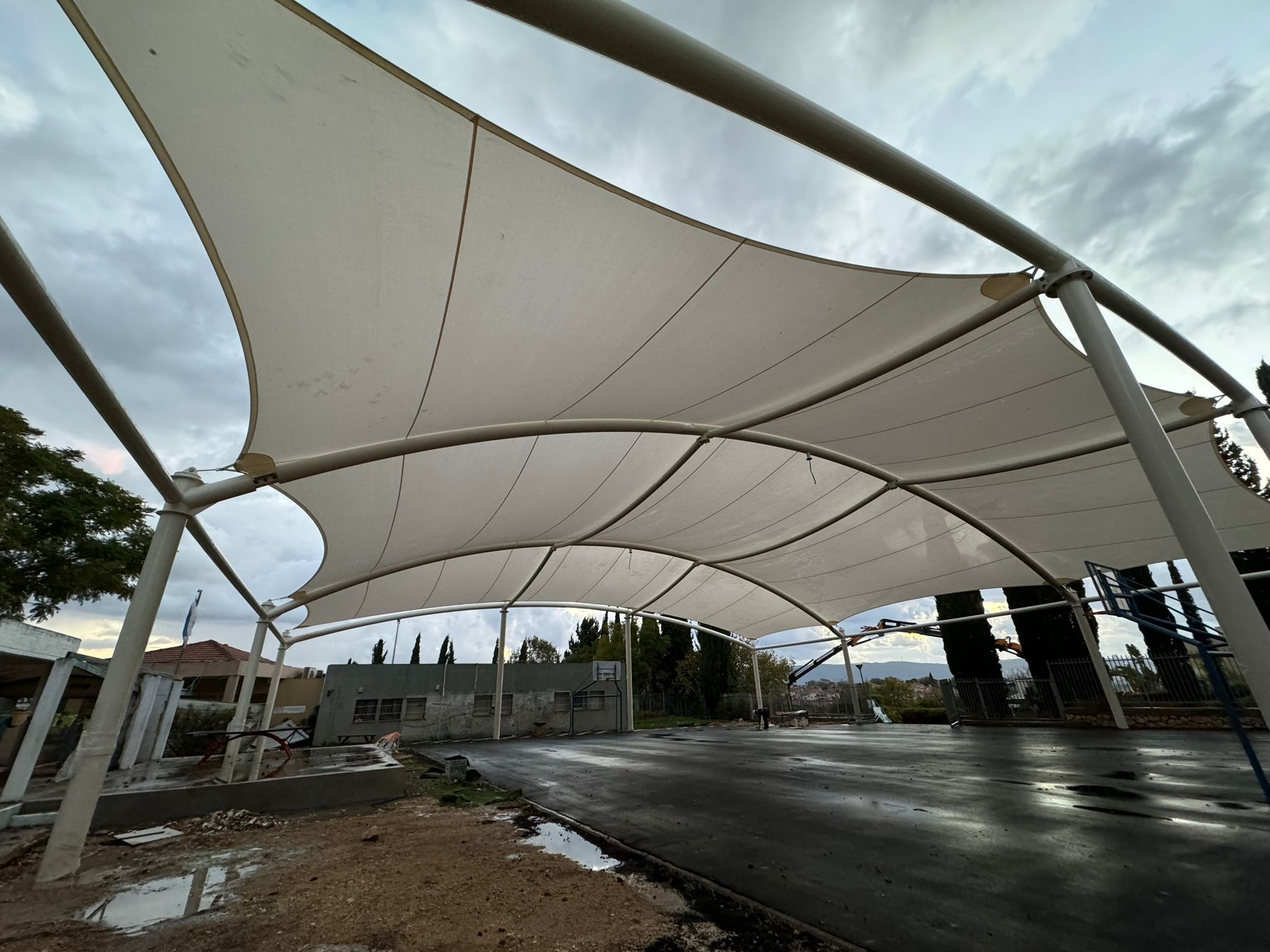 Large beige sunshade with metal frame over paved area under cloudy sky.