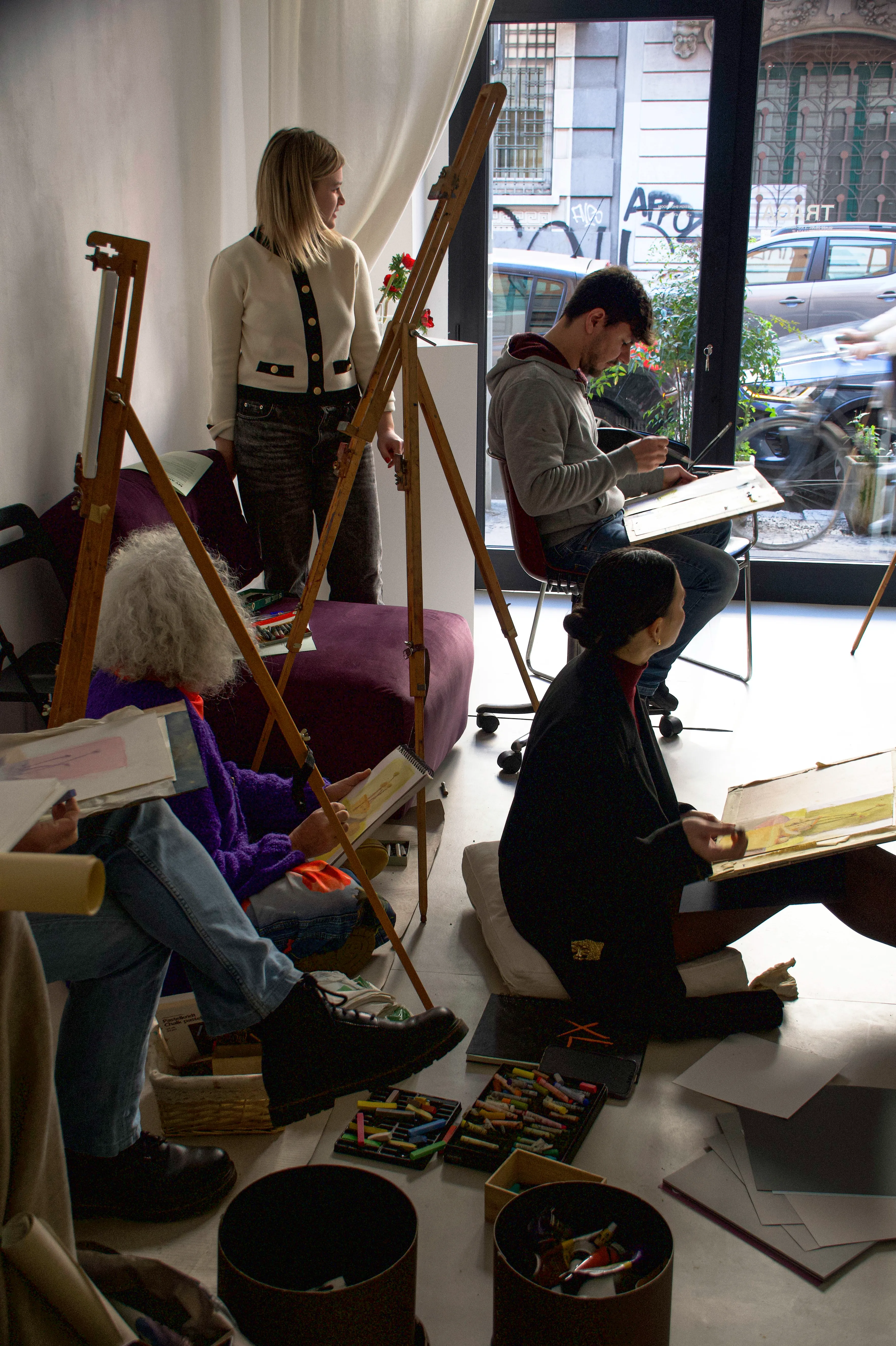 A group of artists are engaged in a life drawing class, seated around wooden easels with sketchpads, in a well-lit studio near a large window overlooking a busy street.