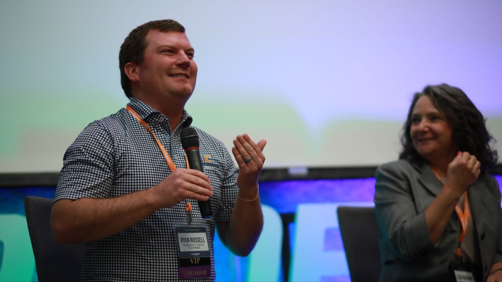 Man with microphone smiles while speaking on stage at PROFCON. His badge reads “Ryan Russell – VIP – Speaker.” A woman next to him also smiles and gestures, both seated in front of a blurred screen backdrop.