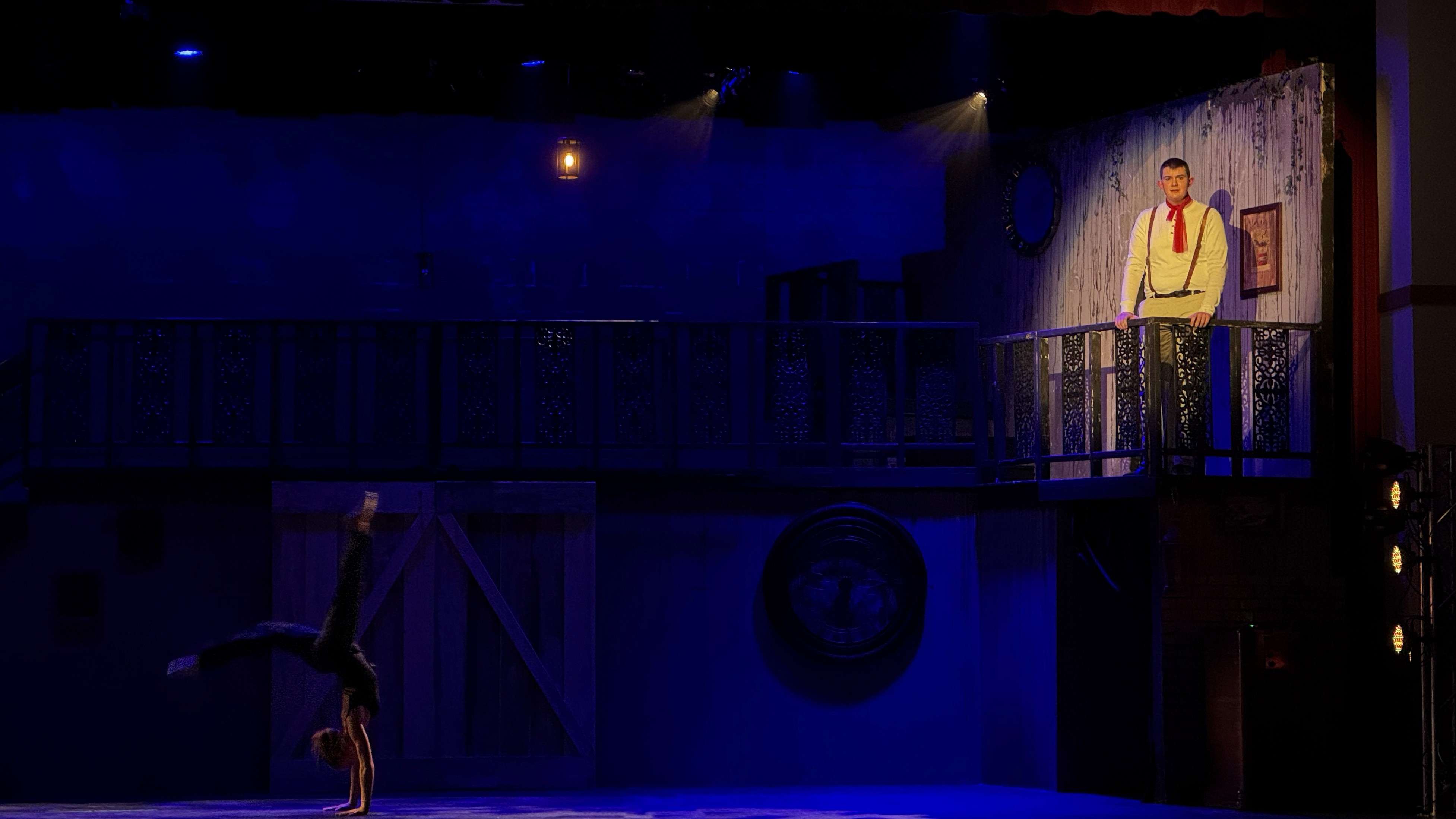 A dancer executes a handstand under warm sidelight isolation and deep blue ambient light, while Orpheus sings Epic II from Hadestown from an upper balcony, creating a dramatic contrast. The image shows side dance lighting and top breakup gobos to light the dancer.