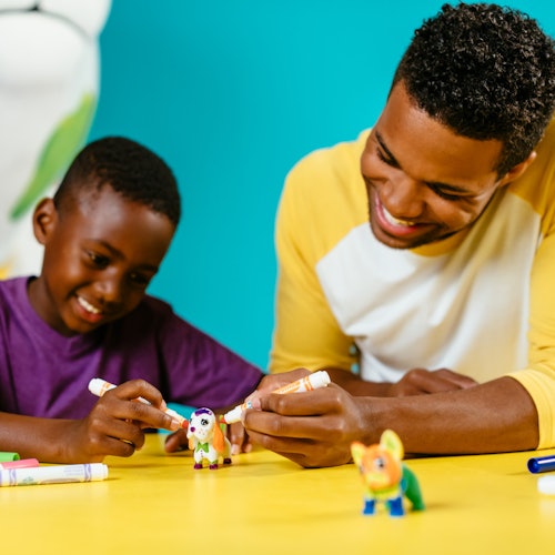 兩個人微笑著在黃色桌子上用記號筆為小玩具 figures 上色，背景是綠松石色。