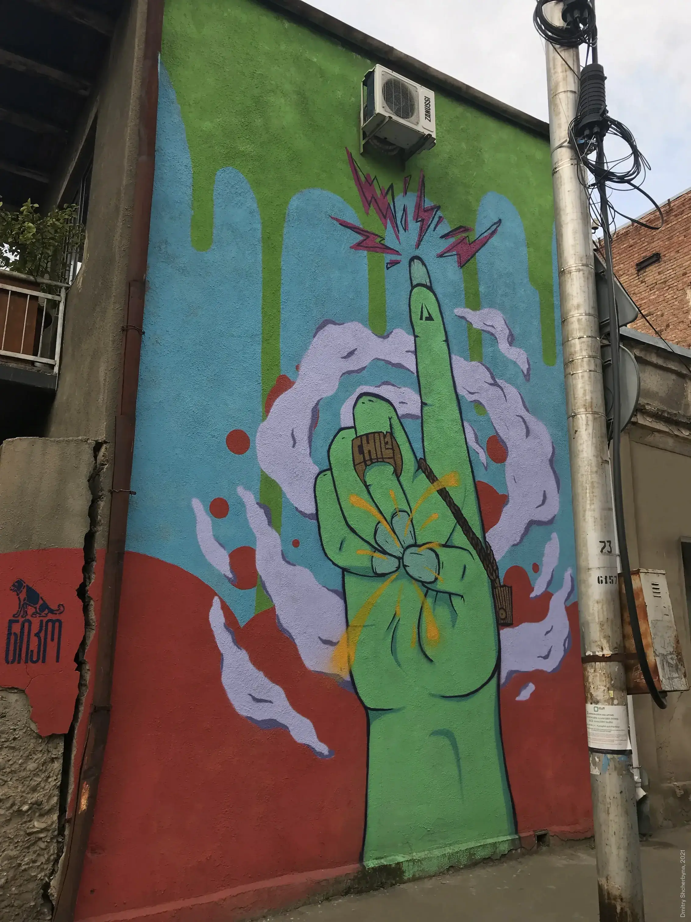 A street mural depicting a large green hand pointing upwards with its fingertip, surrounded by clouds of smoke on a blue and green background. The word "CHILL" is visible on the wrist of the hand.