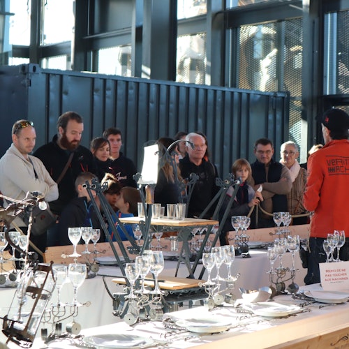 Pessoas reunidas em ambientes fechados, assistindo a uma demonstração com mesas cuidadosamente arrumadas, destacando engenhocas mecânicas e vidrarias.