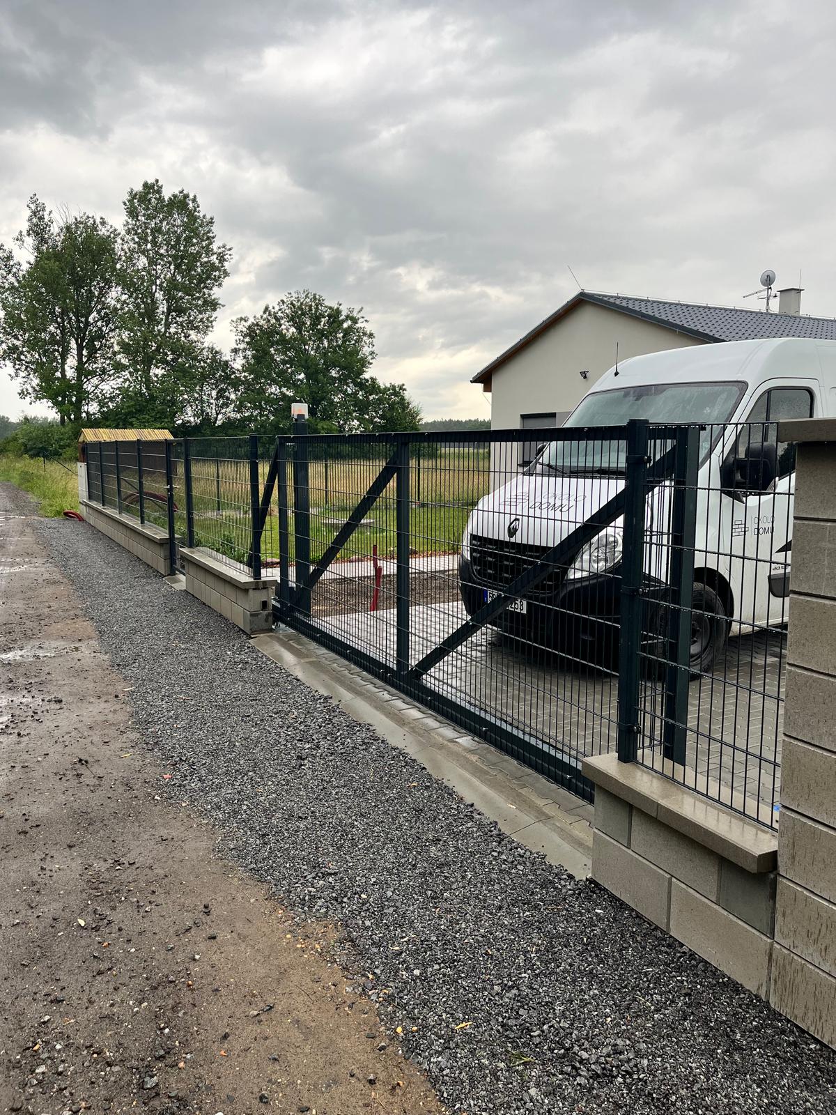 Otevřená kovová brána s černým plotem a zaparkovaným bílým dodávkovým automobilem na štěrkové cestě.