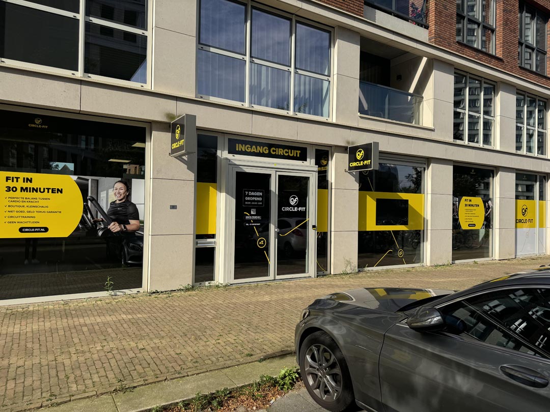 A street view of a Circle Fit hub with large glass doors and yellow banners, alongside a parked car.