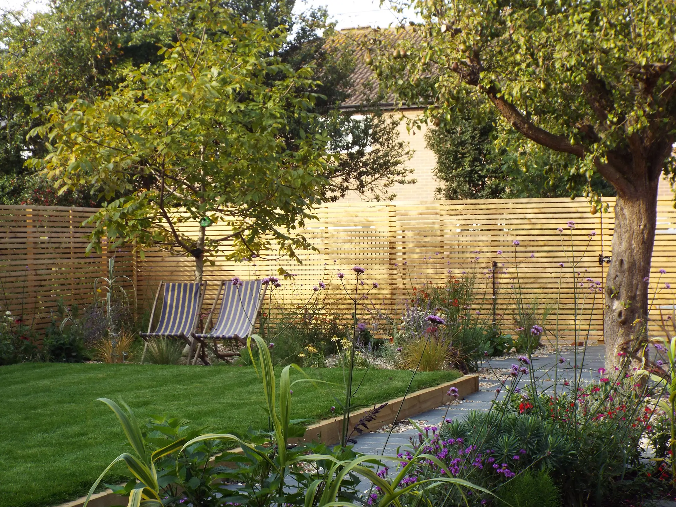 A serene garden scene featuring lush green grass, flowering plants, and a large tree against a wooden fence.