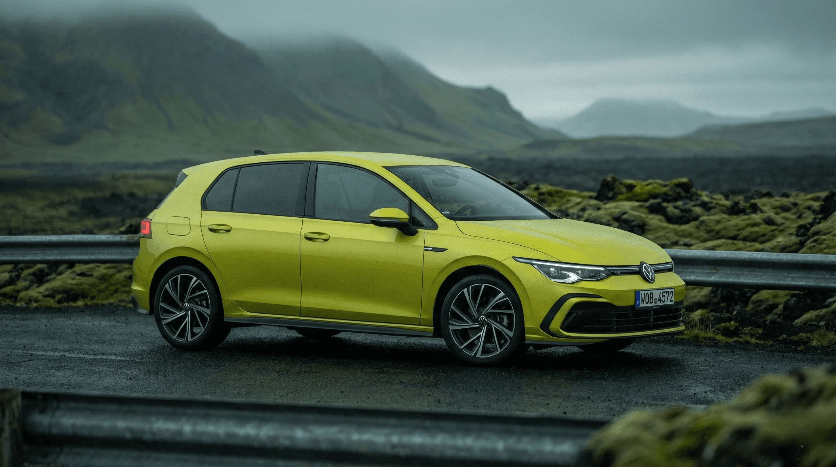 A bright yellow VW Golf parked on a road with green mossy hills and a cloudy sky in the background.