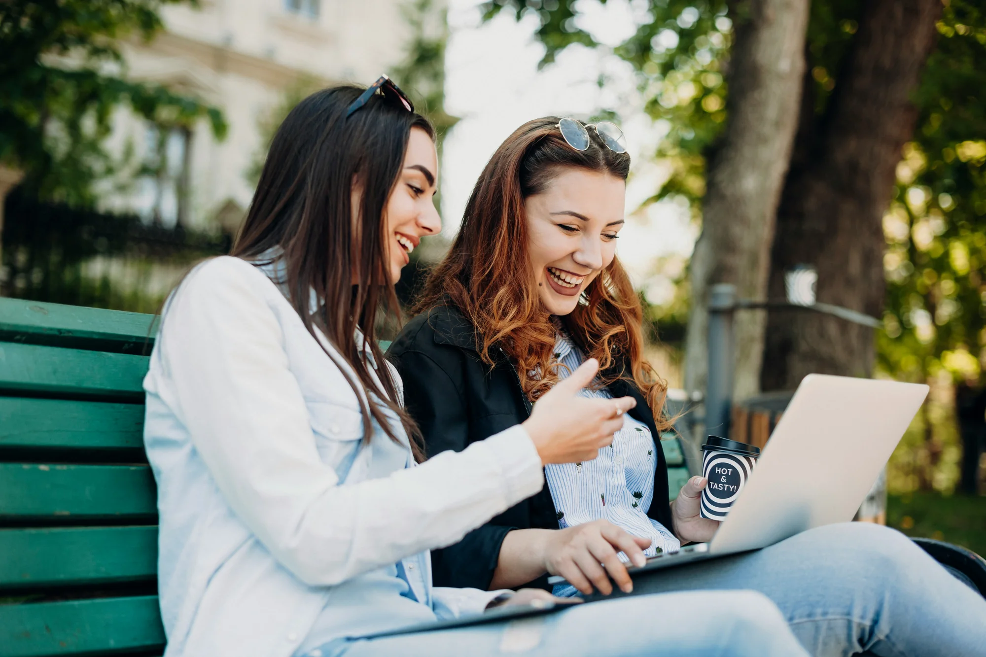 Two women laughing while looking at laptop on park bench outdoors