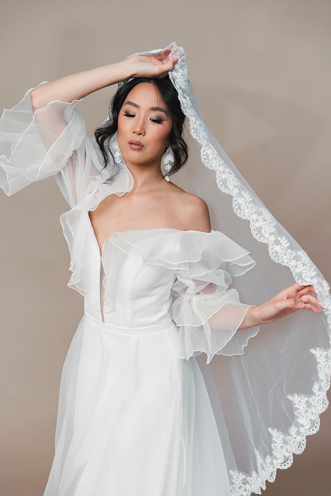 Bride in a gown with sheer lace sleeves and a veil, arm gently raised, fine art bridal editorial by Eva Sitko