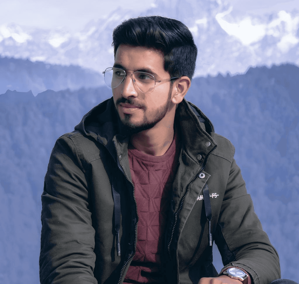 A person stands outdoors in a natural setting with lush green trees under a clear sky, wearing a dark sweatshirt, and looking into the distance.