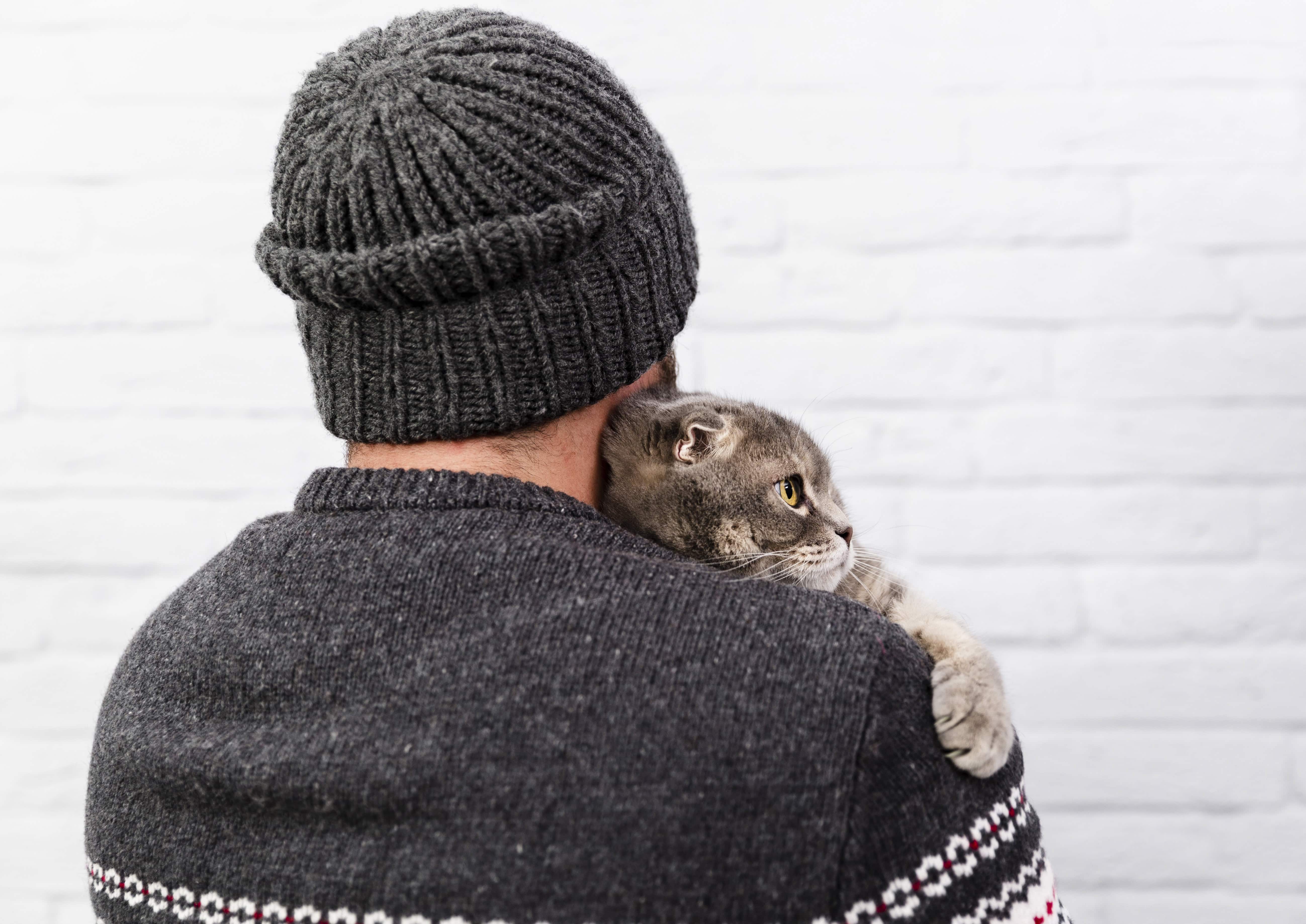 Un homme avec un bonnet gris tient un chat sur son épaule, vue de dos, devant un mur en briques blanches.