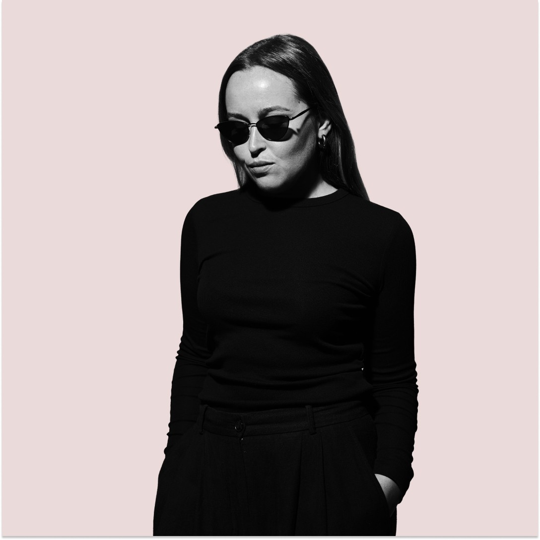 Minimal studio portrait of a confident woman in a white shirt, arms crossed, on a soft gray background.