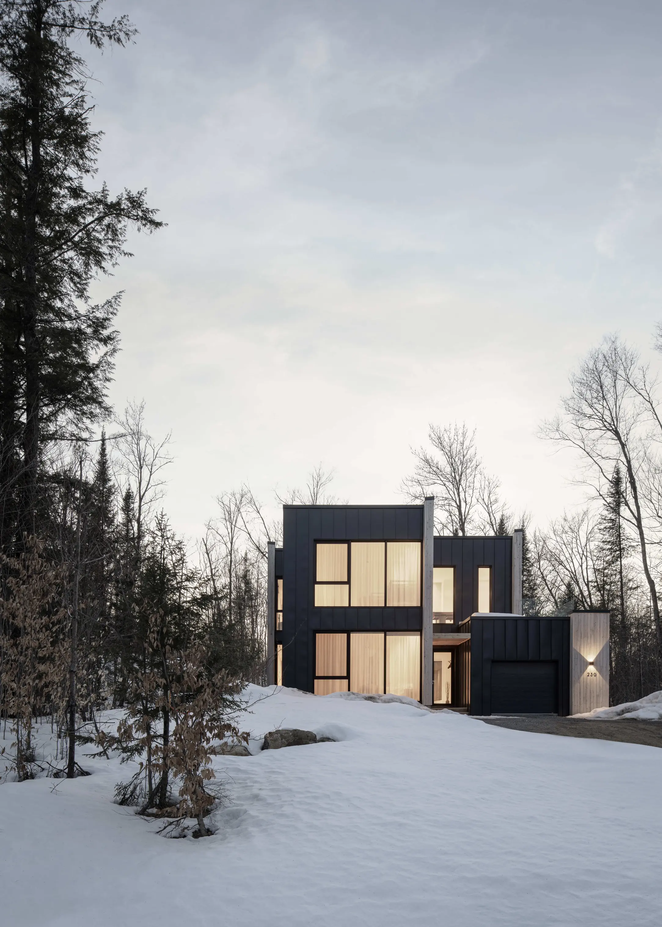 Modern black-clad residence in a snowy forest setting at dusk, with warm interior lighting glowing through large windows.