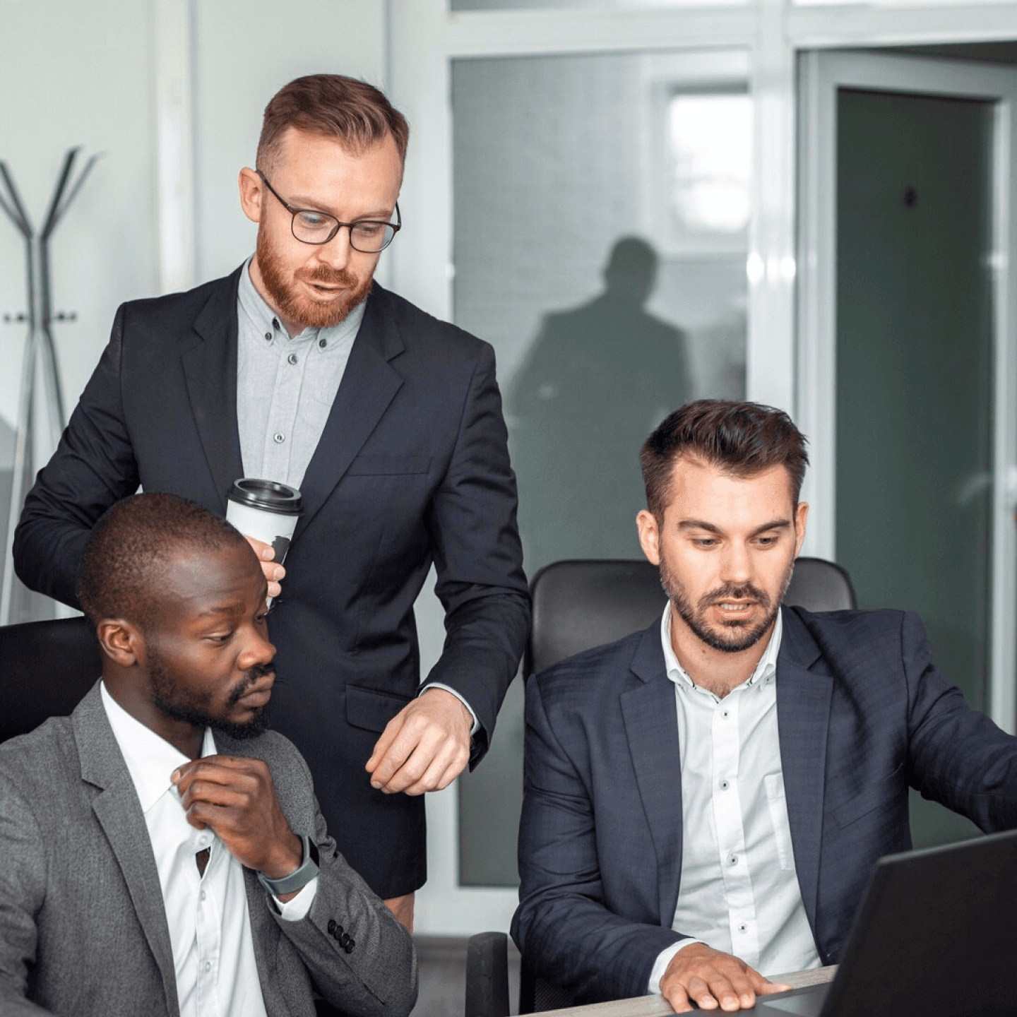 Three people in business attire discussing around a laptop in a modern office.