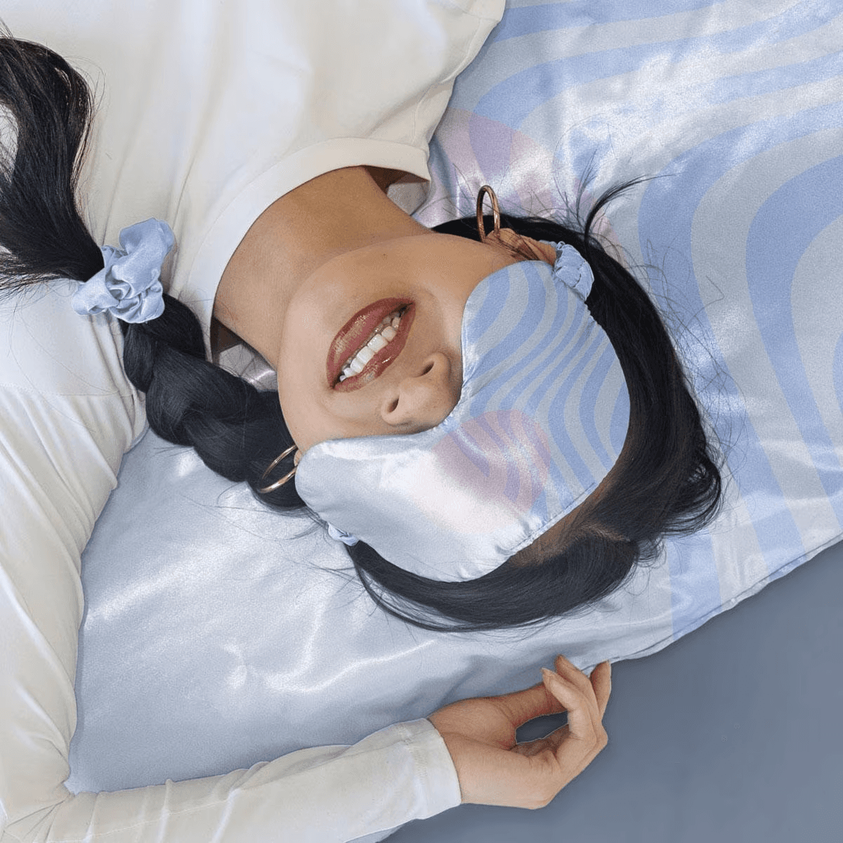 A woman laying down on a synthetic silk pillow case that is light blue and lavendar that has abstract wave patterns across it. She is also wearing an eyemask also made of synthetic silk with the same pattern. Finally her black hair is braided on the side of her head and is held together by a light blue synthetic silk scrunchie.