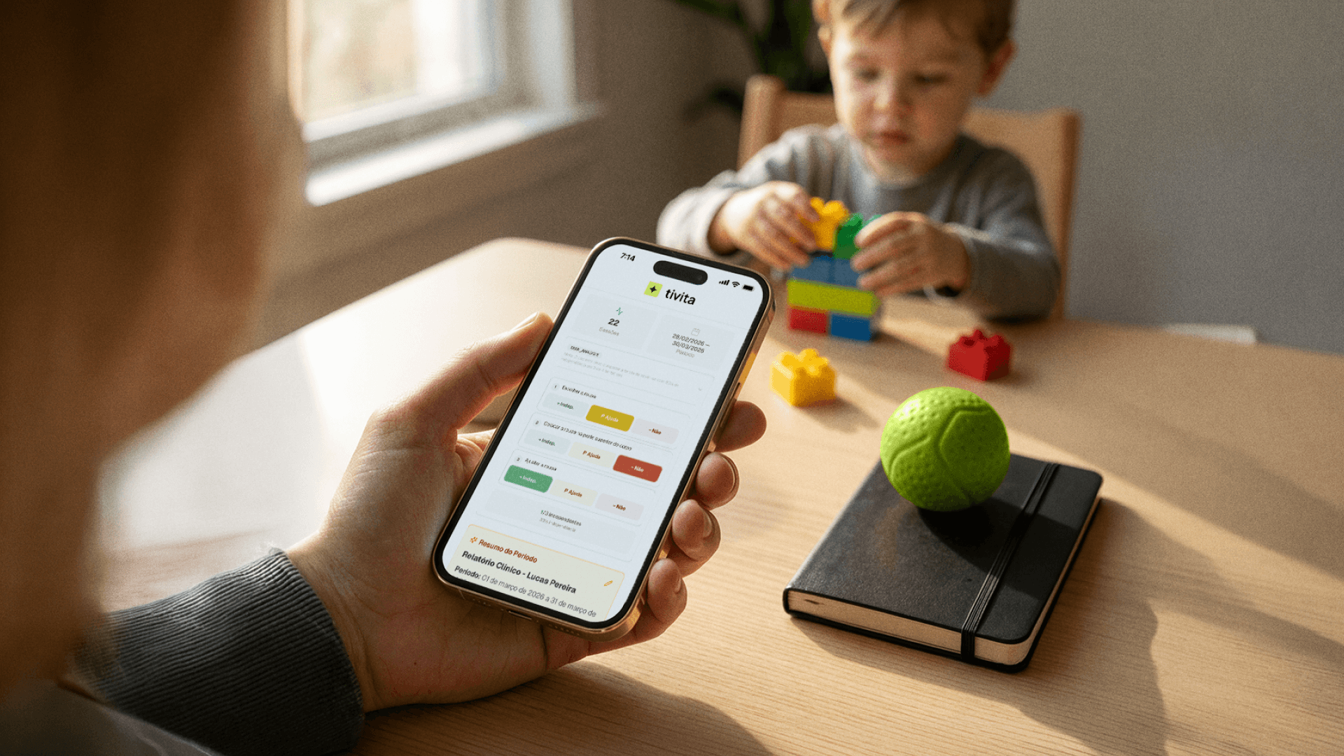 Mão segura um celular exibindo a tela do aplicativo da Tivita com o relatório clínico de uma sessão de terapia, mostrando 22 sessões no período e marcações de desempenho por cores (verde, amarelo e vermelho), enquanto ao fundo uma criança brinca com blocos de montar sobre a mesa, ilustrando o registro de dados de sessão usado por clínicas de terapia ABA na Tivita.