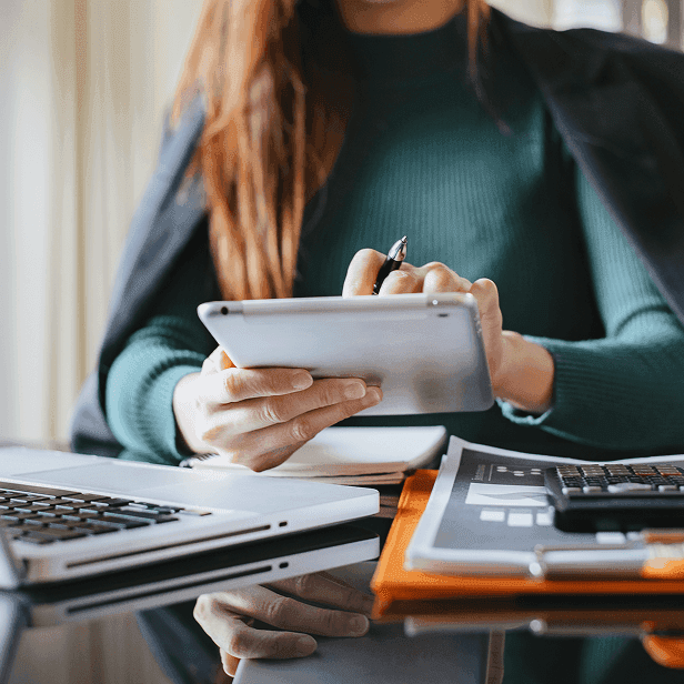 Mulher usando um tablet sobre mesa com notebook, caderno e calculadora.
